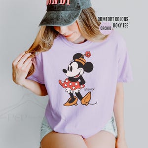 Op de afbeelding: Een lichtpaarse Comfort Colors boxy tee met een Minnie Mouse-afbeelding. De afbeelding toont Minnie Mouse in een rode polkadot rok, oranje schoenen en een bloem in haar haar. Het shirt heeft ook het woord "Disney" erop gedrukt.