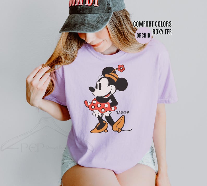 Op de afbeelding: Een lichtpaarse Comfort Colors boxy tee met een Minnie Mouse-afbeelding. De afbeelding toont Minnie Mouse in een rode polkadot rok, oranje schoenen en een bloem in haar haar. Het shirt heeft ook het woord "Disney" erop gedrukt.