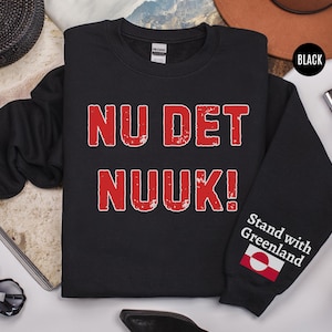 May include: Black sweatshirt with the text "NU DET NUUK!" in red, distressed lettering. The sleeve features the phrase "Stand with Greenland" and a Greenlandic flag. The sweatshirt is a crew neck style.