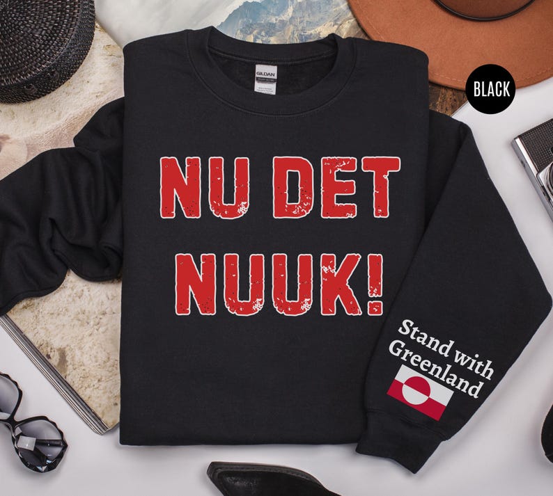 May include: Black sweatshirt with the text "NU DET NUUK!" in red, distressed lettering. The sleeve features the phrase "Stand with Greenland" and a Greenlandic flag. The sweatshirt is a crew neck style.