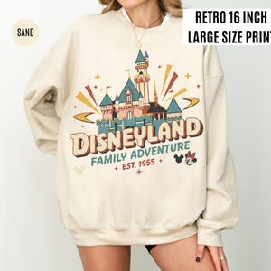 May include: Sand-colored sweatshirt with a retro Disneyland graphic. The design features a castle, the words "DISNEYLAND FAMILY ADVENTURE EST. 1955", and Mickey and Minnie Mouse. The print is 16 inches wide.