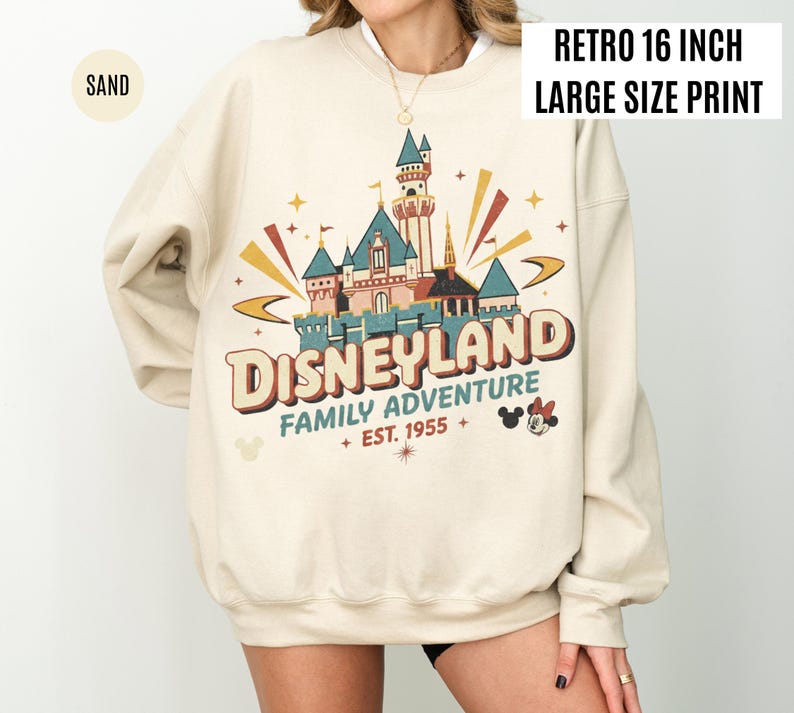 May include: Sand-colored sweatshirt with a retro Disneyland graphic. The design features a castle, the words "DISNEYLAND FAMILY ADVENTURE EST. 1955", and Mickey and Minnie Mouse. The print is 16 inches wide.