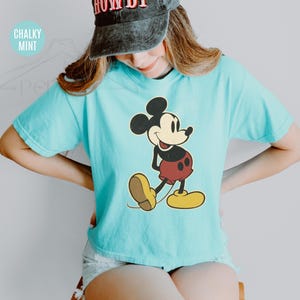 May include: A turquoise t-shirt with a Mickey Mouse design. The cartoon character is in black, red, yellow, and tan. The shirt has short sleeves and a loose fit. The text "Chalky Mint" is in the upper left corner.
