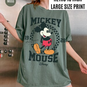May include: A moss green t-shirt with a large Mickey Mouse graphic. The design includes the text "MICKEY MOUSE" and "Disney", along with the numbers "19" and "28". The shirt has a "RETRO 16 INCH LARGE SIZE PRINT" label.