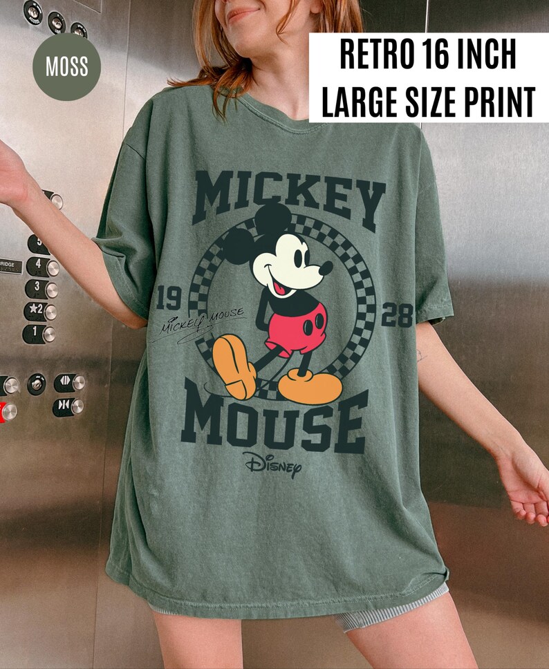 May include: A moss green t-shirt with a large Mickey Mouse graphic. The design includes the text "MICKEY MOUSE" and "Disney", along with the numbers "19" and "28". The shirt has a "RETRO 16 INCH LARGE SIZE PRINT" label.