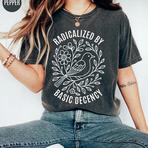 May include: Dark gray t-shirt with the text "RADICALIZED BY BASIC DECENCY" in a white floral design. The design includes a bird, flowers, and leaves. The shirt is a short-sleeved crew neck.