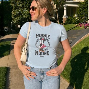 K&ouml;nnte beinhalten: Hellblaues T-Shirt mit einem Minnie Mouse-Motiv in einem karierten Kreis, dem Text "MINNIE MOUSE" und dem Disney-Logo. Das Shirt hat kurze &Auml;rmel und wird mit hellblauen Jeans getragen.