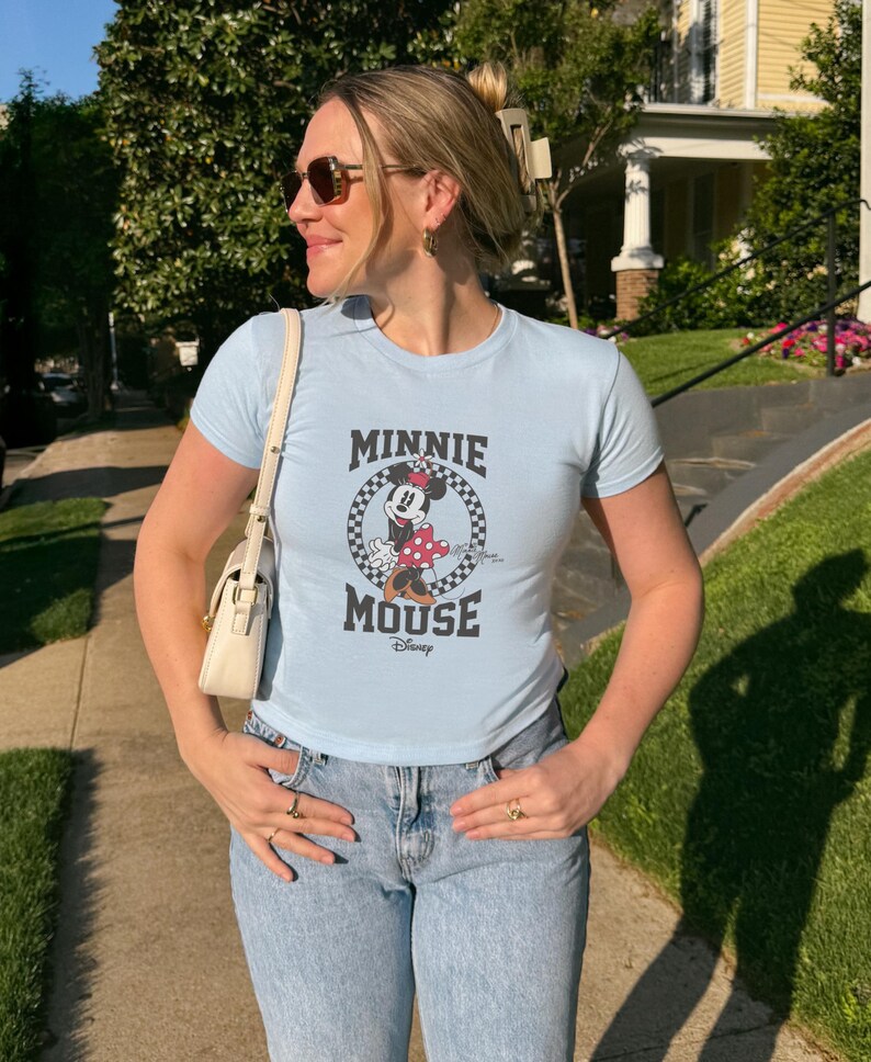 K&ouml;nnte beinhalten: Hellblaues T-Shirt mit einem Minnie Mouse-Motiv in einem karierten Kreis, dem Text "MINNIE MOUSE" und dem Disney-Logo. Das Shirt hat kurze &Auml;rmel und wird mit hellblauen Jeans getragen.