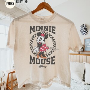 May include: Ivory-colored boxy tee featuring a graphic of Minnie Mouse. The design includes Minnie in a polka-dot dress, set within a checkered circle, with the words "MINNIE MOUSE" above and below the image. The Disney logo is at the bottom.