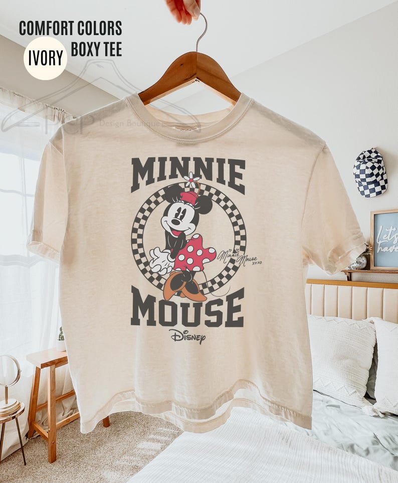 May include: Ivory-colored boxy tee featuring a graphic of Minnie Mouse. The design includes Minnie in a polka-dot dress, set within a checkered circle, with the words "MINNIE MOUSE" above and below the image. The Disney logo is at the bottom.