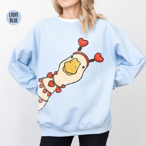 Pode incluir: Camisola azul clara com uma ilustra&ccedil;&atilde;o l&uacute;dica de um ganso adornado com detalhes de cora&ccedil;&atilde;o vermelho. O ganso usa uma faixa de cabe&ccedil;a em forma de cora&ccedil;&atilde;o e um colar de cora&ccedil;&otilde;es. O texto "LIGHT BLUE" est&aacute; em um c&iacute;rculo azul claro no canto superior esquerdo.