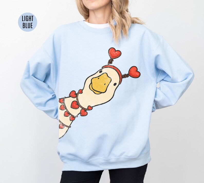 Pode incluir: Camisola azul clara com uma ilustra&ccedil;&atilde;o l&uacute;dica de um ganso adornado com detalhes de cora&ccedil;&atilde;o vermelho. O ganso usa uma faixa de cabe&ccedil;a em forma de cora&ccedil;&atilde;o e um colar de cora&ccedil;&otilde;es. O texto "LIGHT BLUE" est&aacute; em um c&iacute;rculo azul claro no canto superior esquerdo.