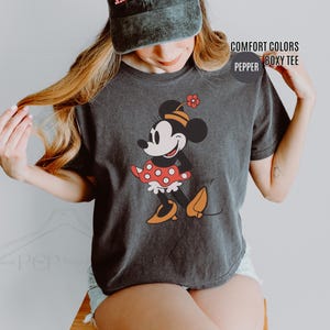 Op de afbeelding: Donkergrijze Comfort Colors boxy tee met een Minnie Mouse-afbeelding. Het stripfiguur draagt een rood-witte polkadotjurk, oranje schoenen en een hoed met een bloem. Op het shirt staan de woorden "COMFORT COLORS" en "PEPPER".
