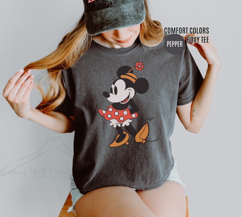 Op de afbeelding: Donkergrijze Comfort Colors boxy tee met een Minnie Mouse-afbeelding. Het stripfiguur draagt een rood-witte polkadotjurk, oranje schoenen en een hoed met een bloem. Op het shirt staan de woorden "COMFORT COLORS" en "PEPPER".