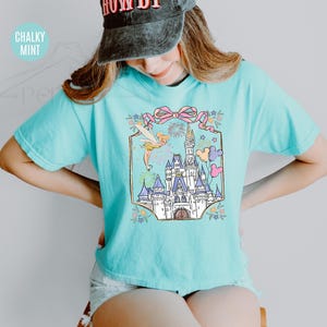 May include: A light blue t-shirt featuring a colorful graphic of a fairytale castle, Tinkerbell, fireworks, and balloons. The graphic is framed by a pink bow and floral accents. The shirt is a comfortable, casual style.