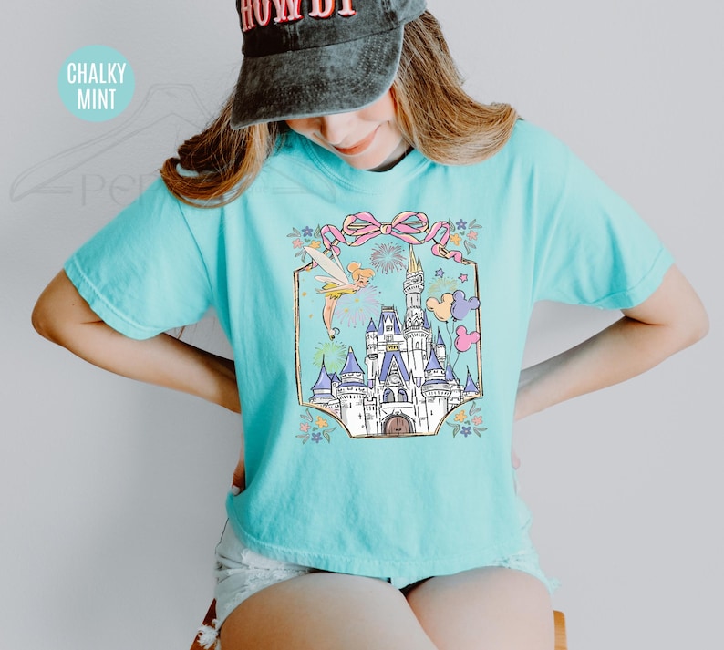 May include: A light blue t-shirt featuring a colorful graphic of a fairytale castle, Tinkerbell, fireworks, and balloons. The graphic is framed by a pink bow and floral accents. The shirt is a comfortable, casual style.