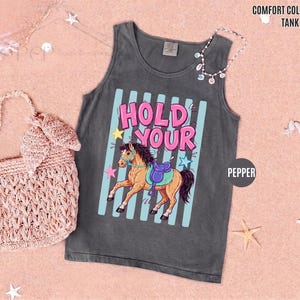 K&ouml;nnte beinhalten: Ein graues Tanktop mit den Worten "HOLD YOUR" in Pink und Blau sowie einer Cartoon-Pferdeillustration. Das Tanktop geh&ouml;rt zur "Comfort Colors"-Kollektion und ist mit "Pepper" gekennzeichnet. Eine rosa gewebte Tasche und eine Halskette sind ebenfalls im Bild.
