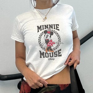 K&ouml;nnte beinhalten: Wei&szlig;es T-Shirt mit einem Minnie Mouse-Motiv in einem schwarz-wei&szlig; karierten Kreis und dem Schriftzug "MINNIE MOUSE" in Schwarz. Das Disney-Logo befindet sich darunter. Das T-Shirt hat kurze &Auml;rmel.