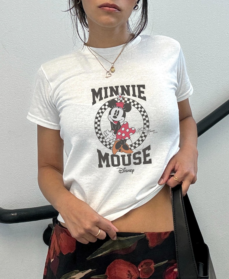 K&ouml;nnte beinhalten: Wei&szlig;es T-Shirt mit einem Minnie Mouse-Motiv in einem schwarz-wei&szlig; karierten Kreis und dem Schriftzug "MINNIE MOUSE" in Schwarz. Das Disney-Logo befindet sich darunter. Das T-Shirt hat kurze &Auml;rmel.