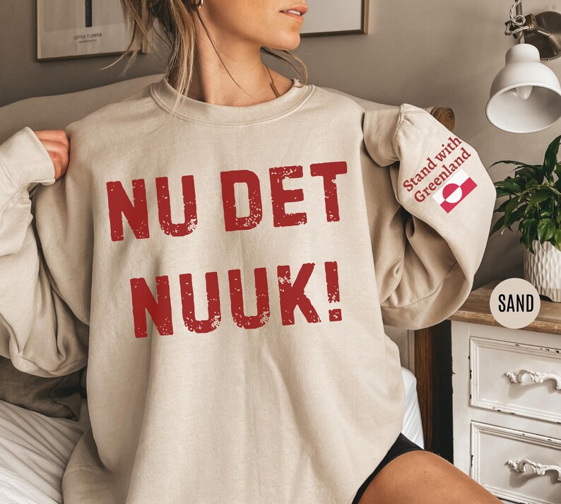 May include: Sand-colored sweatshirt with the text "NU DET NUUK!" in distressed red lettering. The sleeve features a "Stand with Greenland" graphic and the word "SAND" in a circle.