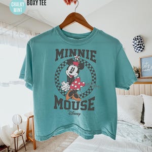 May include: A mint green, short-sleeved t-shirt featuring a graphic of Minnie Mouse. The design includes the words "MINNIE MOUSE" in a dark gray font, with Minnie in a checkered circle. The shirt is hanging on a wooden hanger.