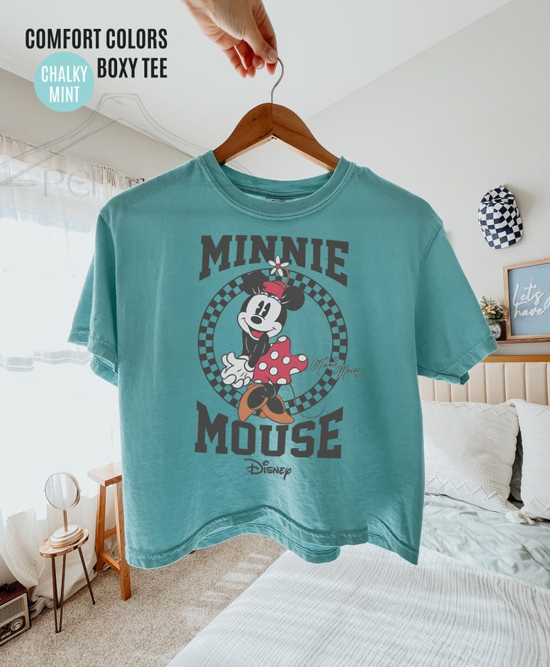 May include: A mint green, short-sleeved t-shirt featuring a graphic of Minnie Mouse. The design includes the words "MINNIE MOUSE" in a dark gray font, with Minnie in a checkered circle. The shirt is hanging on a wooden hanger.