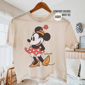 Op de afbeelding: Ivoor boxy T-shirt met een Minnie Mouse-afbeelding. Het stripfiguur wordt afgebeeld in een klassieke pose, met een rode polkadot rok, oranje schoenen en een bijpassende bloemenhoed. Het woord "Disney" staat eronder gedrukt.