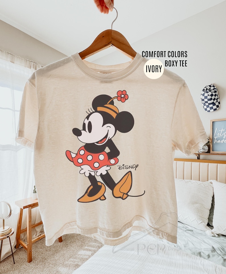 Op de afbeelding: Ivoor boxy T-shirt met een Minnie Mouse-afbeelding. Het stripfiguur wordt afgebeeld in een klassieke pose, met een rode polkadot rok, oranje schoenen en een bijpassende bloemenhoed. Het woord "Disney" staat eronder gedrukt.