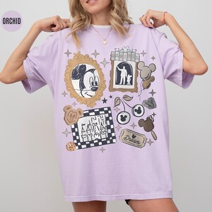 May include: Orchid-colored t-shirt featuring a collage of Disney-themed graphics. The design includes Mickey Mouse, a castle, cherries, and other elements. The shirt has a relaxed fit and short sleeves.
