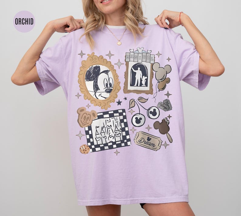 May include: Orchid-colored t-shirt featuring a collage of Disney-themed graphics. The design includes Mickey Mouse, a castle, cherries, and other elements. The shirt has a relaxed fit and short sleeves.