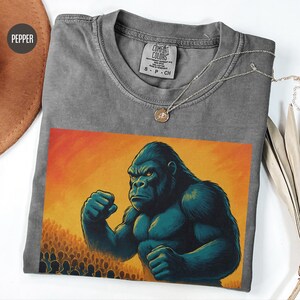 May include: A gray t-shirt with a vintage wash featuring a graphic of a large gorilla with its fists clenched, standing in front of a crowd of people. The text "COMFORT COLORS" is on the tag.