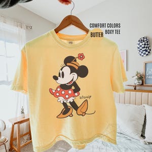 Op de afbeelding: Een gele Comfort Colors boxy tee met een Minnie Mouse-afbeelding. Het stripfiguur draagt een rode polkadot rok, oranje schoenen en een bloem in haar haar. Het Disney-logo staat eronder gedrukt.
