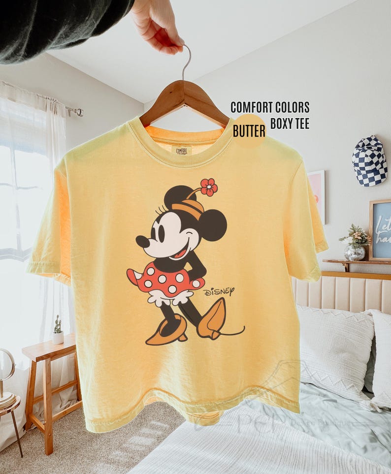 Op de afbeelding: Een gele Comfort Colors boxy tee met een Minnie Mouse-afbeelding. Het stripfiguur draagt een rode polkadot rok, oranje schoenen en een bloem in haar haar. Het Disney-logo staat eronder gedrukt.