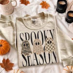 May include: A beige crewneck sweatshirt featuring the black text SPOOKY SEASON and three ghost illustrations. The ghosts have different patterns: one in camouflage, one in leopard print, and one in a tan and white checkerboard design.