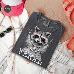 May include: Dark grey t-shirt featuring a raccoon wearing pink sunglasses and a pearl necklace, with the word "FERAL" in white. The raccoon is making a rock and roll hand gesture. The shirt is folded on a white and grey striped fabric.