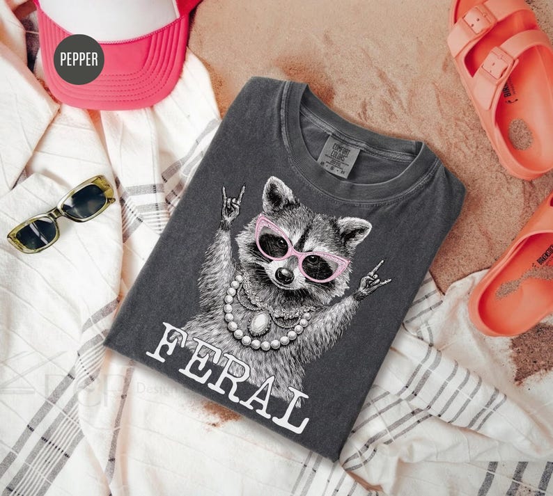 May include: Dark grey t-shirt featuring a raccoon wearing pink sunglasses and a pearl necklace, with the word "FERAL" in white. The raccoon is making a rock and roll hand gesture. The shirt is folded on a white and grey striped fabric.