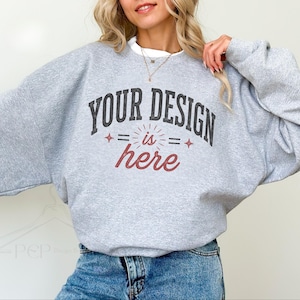 May include: A heather gray crewneck sweatshirt featuring the dark gray text YOUR DESIGN and the red cursive text is here, accented by small red star symbols and horizontal lines, worn over a white shirt with blue denim jeans.