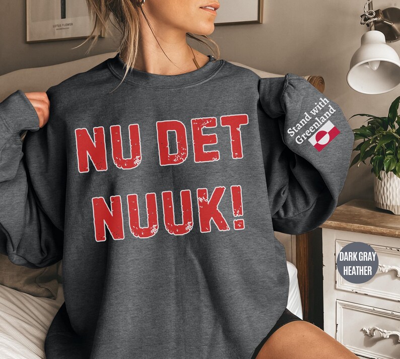 May include: Dark gray heather sweatshirt with the text "NU DET NUUK!" in distressed red lettering. The right sleeve features the text "Stand with Greenland" and a Greenlandic flag. The sweatshirt is a casual style.