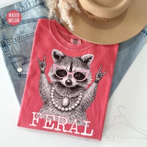 May include: A coral-colored t-shirt featuring a raccoon wearing pink sunglasses and a pearl necklace, with the word "FERAL" in white block letters. The shirt is laid on top of blue jeans and a tan hat.