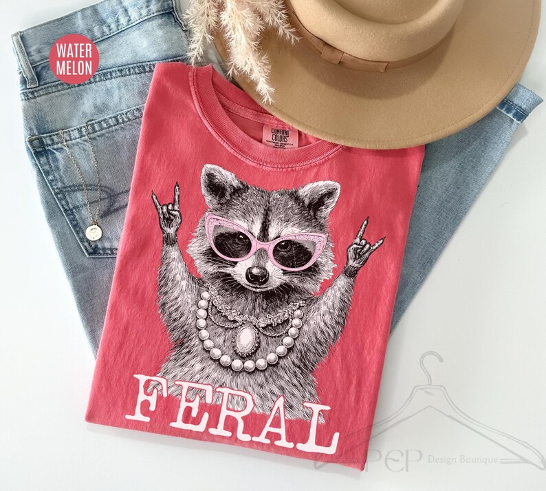May include: A coral-colored t-shirt featuring a raccoon wearing pink sunglasses and a pearl necklace, with the word "FERAL" in white block letters. The shirt is laid on top of blue jeans and a tan hat.