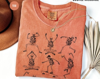 May include: Coral-colored t-shirt with a crew neck and short sleeves. The shirt features black skeleton illustrations in various poses, each with the text "FUCK TRUMP" incorporated into the design. A gold necklace with a pendant is draped on the shirt.