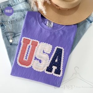 May include: A violet t-shirt featuring the letters USA in a decorative applique style. The U is red and white striped, the S is white with a subtle plaid pattern, and the A is navy blue with white polka dots. Each letter has a white scalloped trim.