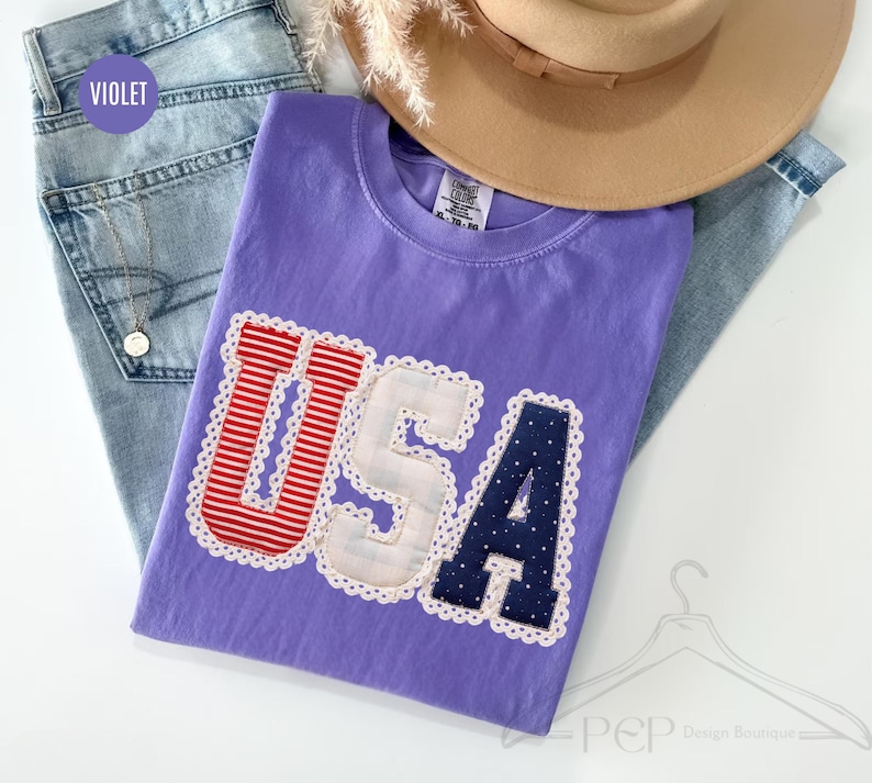 May include: A violet t-shirt featuring the letters USA in a decorative applique style. The U is red and white striped, the S is white with a subtle plaid pattern, and the A is navy blue with white polka dots. Each letter has a white scalloped trim.