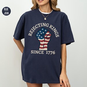 Puede incluir: Camiseta azul marino con el texto "Rejecting Kings Since 1776" que rodea un gr&aacute;fico de un pu&ntilde;o con los colores de la bandera estadounidense. La camiseta tiene mangas cortas y cuello redondo.