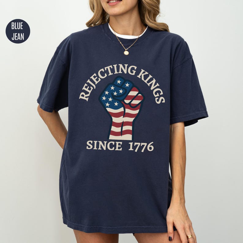 Puede incluir: Camiseta azul marino con el texto "Rejecting Kings Since 1776" que rodea un gr&aacute;fico de un pu&ntilde;o con los colores de la bandera estadounidense. La camiseta tiene mangas cortas y cuello redondo.