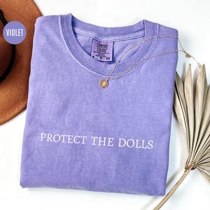 May include: A purple t-shirt with the text "PROTECT THE DOLLS" printed in white on the front.