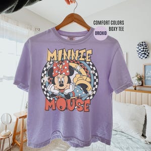 Może przedstawiać: Fioletowa koszulka Comfort Colors boxy z grafiką Myszki Minnie. Projekt zawiera twarz Minnie, czerwoną kokardę oraz napis "MINNIE MOUSE" w stylu retro. Koszulka wisi na drewnianym wieszaku.