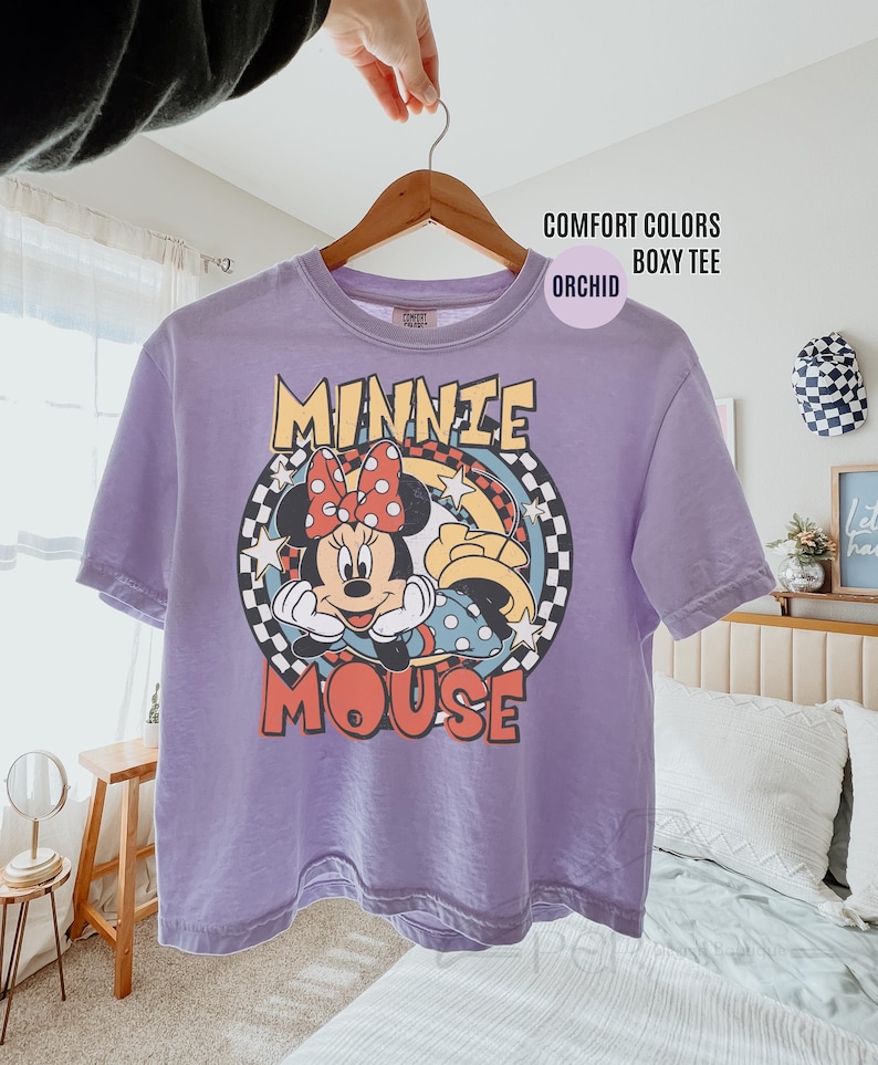 Może przedstawiać: Fioletowa koszulka Comfort Colors boxy z grafiką Myszki Minnie. Projekt zawiera twarz Minnie, czerwoną kokardę oraz napis "MINNIE MOUSE" w stylu retro. Koszulka wisi na drewnianym wieszaku.