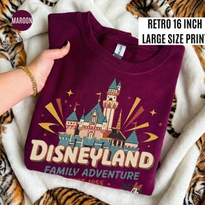 May include: Maroon crewneck sweatshirt with a Disneyland graphic. The design features a castle, stars, and the words "Disneyland Family Adventure Est. 1955." The print is 16 inches in size.