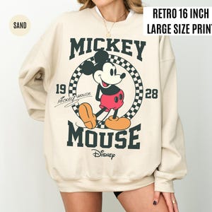 Może przedstawiać: Beżowa bluza z dużym nadrukiem Myszki Miki. Projekt zawiera tekst "Mickey Mouse" i "Disney" z rokiem "1928". Bluza ma nadruk w stylu retro o wielkości 40,6 cm.